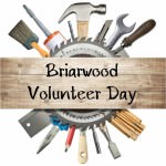 An unfinished plank for wood with the words "Briarwood Volunteer Day" in the foreground; behind the plank is a circular saw and a round array of construction tools.