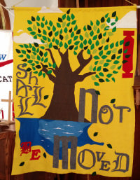 [Image description: Photo of a church banner showing a tree with a brown trunk and green leaves, planted near water; the banner background is yellow, and the words "I shall not be moved" are written around hte image in red and gray text.]