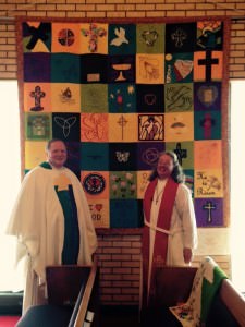 [Image description: Bishop Kevin Kanouse and Pastor Bonita Knox, wearing pastor's robes, stand in front of a colorful handmade quilt, made of squares in shades of yellow and green, with various images on them.]