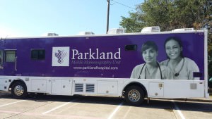 [Image description: photo of a Parkland Hospital mobile health center bus. The bus is decorated with a deep purple background and two female doctors or nurses are picture on the right side of the bus in black and white. The Parkland logo and name appear next to the women's faces. The bus is parked in a parking lot on a sunny day.]