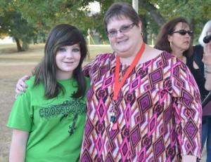 [Image description: Two white women, one a teen with long brown hair and wearing a green Crossroads t-shirt, the other an adult woman with short brown hair and glasses, wearing a patterned purple and pink shirt, stand outdoors, facing the camera and smiling. The older woman has her arm around the teen.]