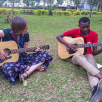 [Image description: A young white woman sits cross-legged on the grass, leaning over her guitar. To her left is a young Black man sitting on the grass with his legs out in front of him, his ankles crossed. He is also playing a guitar.]