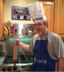 [Image desceription: a white man with gray hair and a beard stands in front of a stove. He is wearing a white chef's hat reading "Lefse Maker" and a blue apron that also says "Lefse." He is holding up a rolling pin.]