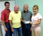 [Image description: four people stand facing the camera, smiling, three men on the left and a woman on the right. The men are presenting a check to the woman.]