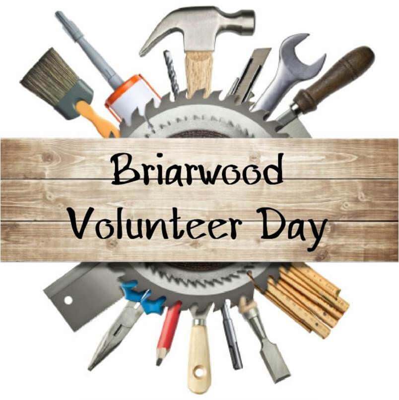 An unfinished plank for wood with the words "Briarwood Volunteer Day" in the foreground; behind the plank is a circular saw and a round array of construction tools.